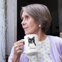 A woman holds a Domino cat portrait mug beside a window.