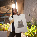 A woman holds a personalized gray cat poster in a sunlit courtyard.